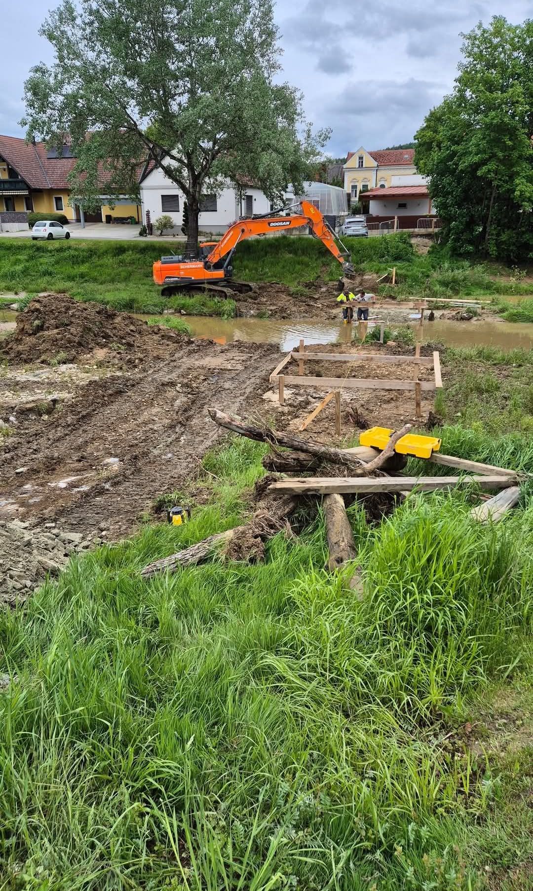 Baustelle mit einem orangefarbenen Bagger, zwei Arbeitern in gelben Westen, Holzkonstruktionen und Erdhaufen.