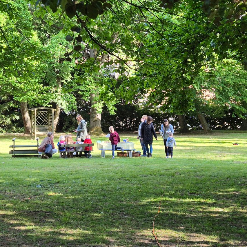Eine Gruppe von Menschen versammelt sich in einem Park. Einige sitzen auf Bänken, während andere herumlaufen. Ein kleines Kind spielt auf dem Gras. Im Hintergrund befinden sich Bäume und ein Schuppen.