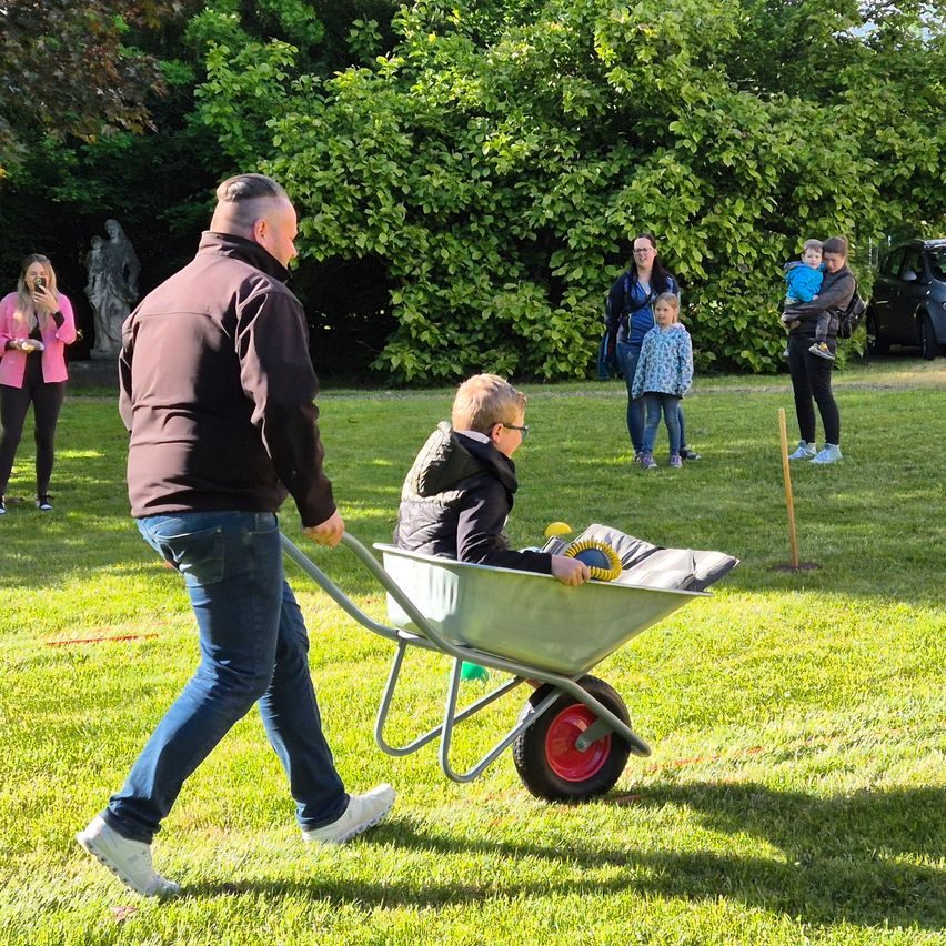 Ein Mann schiebt eine Schubkarre mit einem Jungen darin in einem Park, wo mehrere Leute zuschauen.