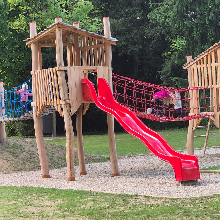 Eine Holzspielanlage mit einer roten Rutsche und einem Tauseil. Kinder klettern auf der Anlage.