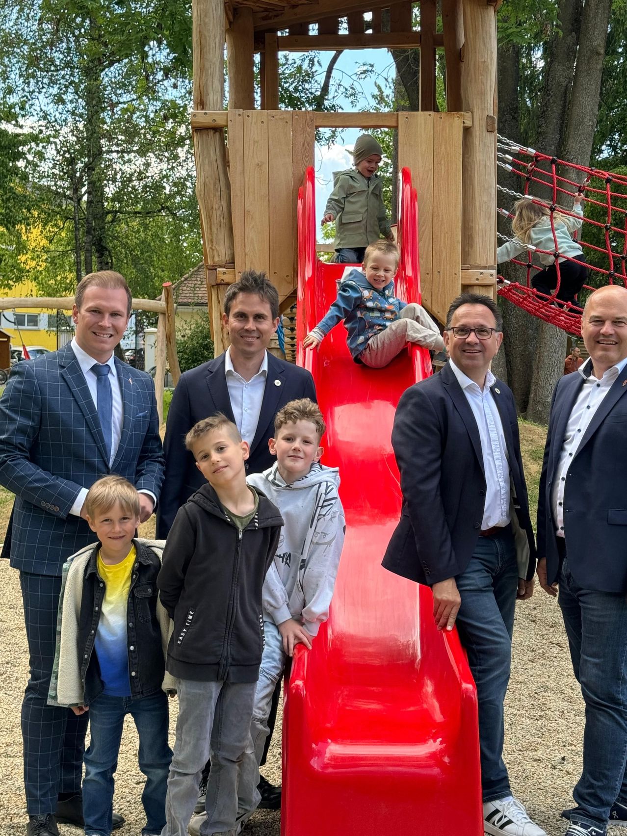 Eine Gruppe von Erwachsenen und Kindern posiert für ein Foto im Park. Zwei Kinder spielen auf der roten Rutsche, während andere in der Nähe stehen.