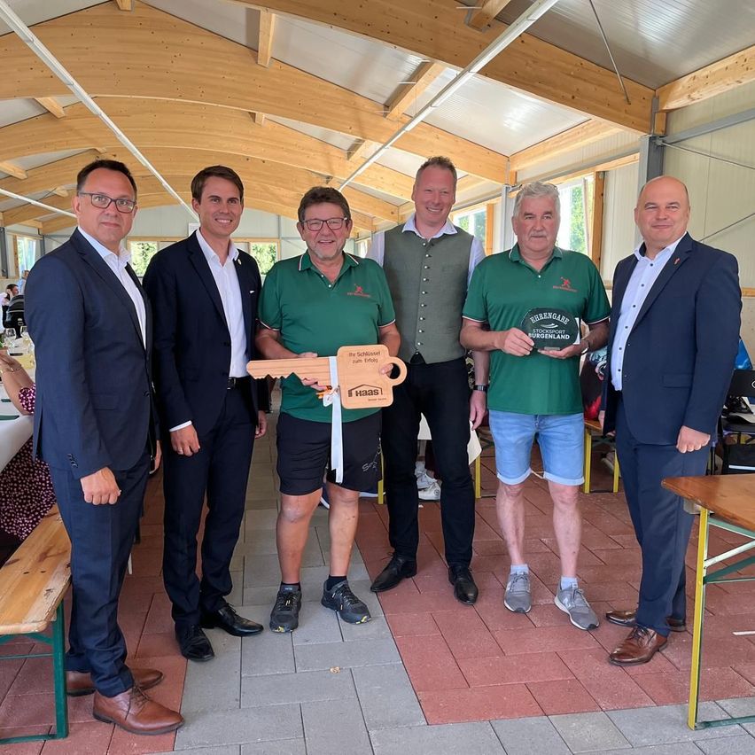 Fünf Männer stehen drinnen, möglicherweise in einem Clubhaus. Zwei Männer in Grün halten eine Trophäe und einen Schlüssel, während andere lächeln. Dahinter ist eine Holzkonstruktion mit Fenstern und einem Tisch zu sehen.