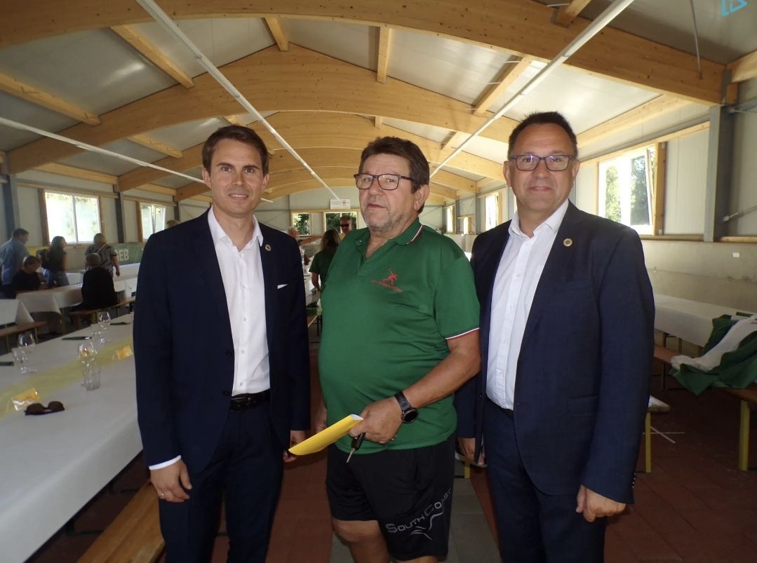 Drei Männer stehen zusammen in einem Saal. Ein Mann in Grün trägt eine Brille und hält eine Mappe. Der Mann zu seiner Linken trägt einen schwarzen Anzug, und der Mann zu seiner Rechten trägt einen dunkelblauen Anzug. Hinter ihnen sind Tische mit Weingläsern gedeckt.