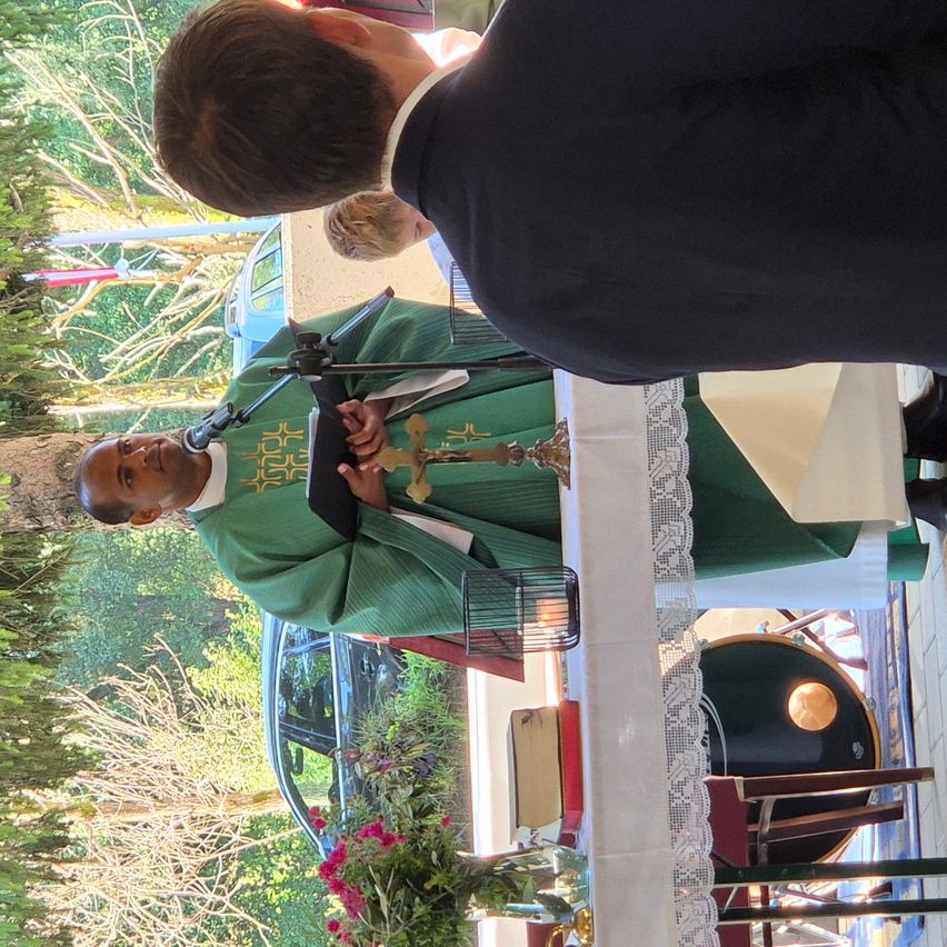 Ein Priester in grünen Gewändern steht an einem Rednerpult und hält eine Rede. Ein Mikrofon und ein Kreuz sind vor ihm platziert. Eine weitere Person steht hinter ihm, und ein Auto ist auf der linken Seite geparkt.