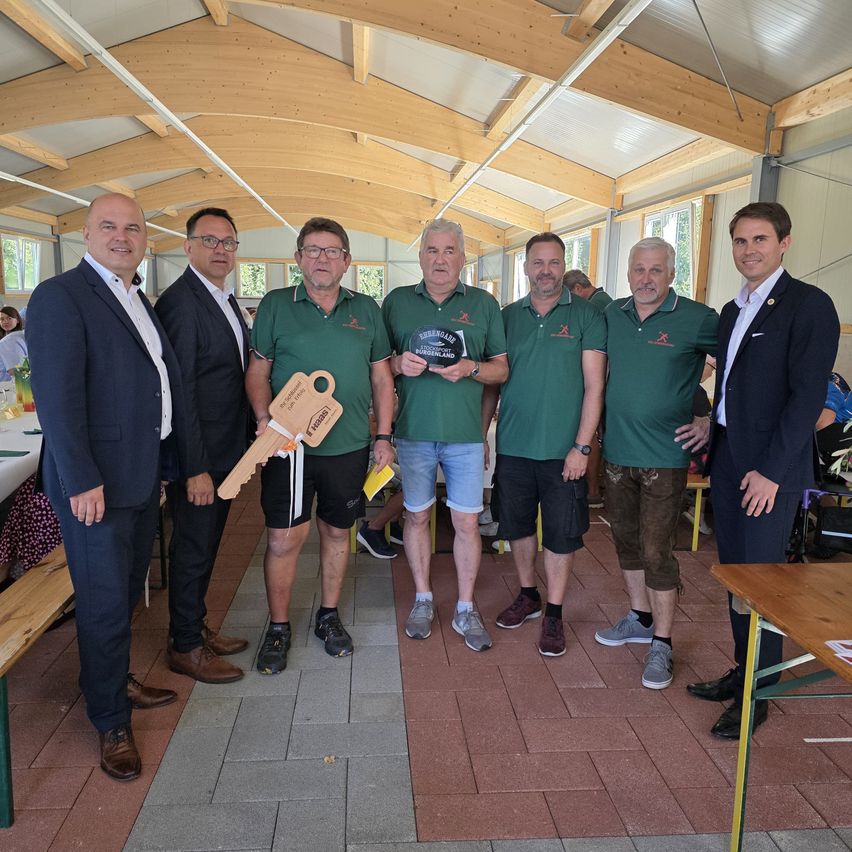Sieben Männer stehen in einem Raum und halten eine Trophäe und einen Schlüssel. Sie tragen alle grüne Poloshirts. Der Raum hat Holzbalken und Fenster.