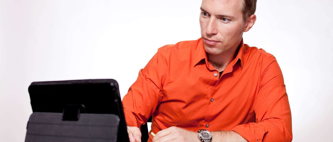 Ein Mann in einem orangefarbenen Hemd sitzt am Schreibtisch und schreibt mit einem Stift. Er hat ein Tablet und ein offenes Buch auf dem Schreibtisch.