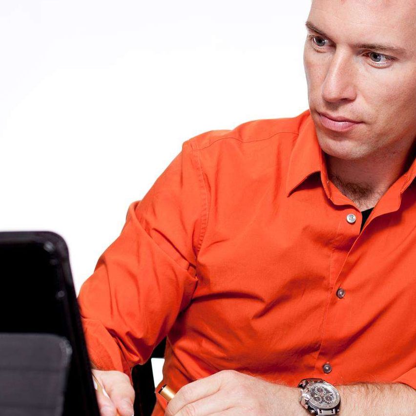 Ein Mann in einem orangefarbenen Hemd sitzt am Schreibtisch und schreibt mit einem Stift. Er hat ein Tablet und ein offenes Buch auf dem Schreibtisch.