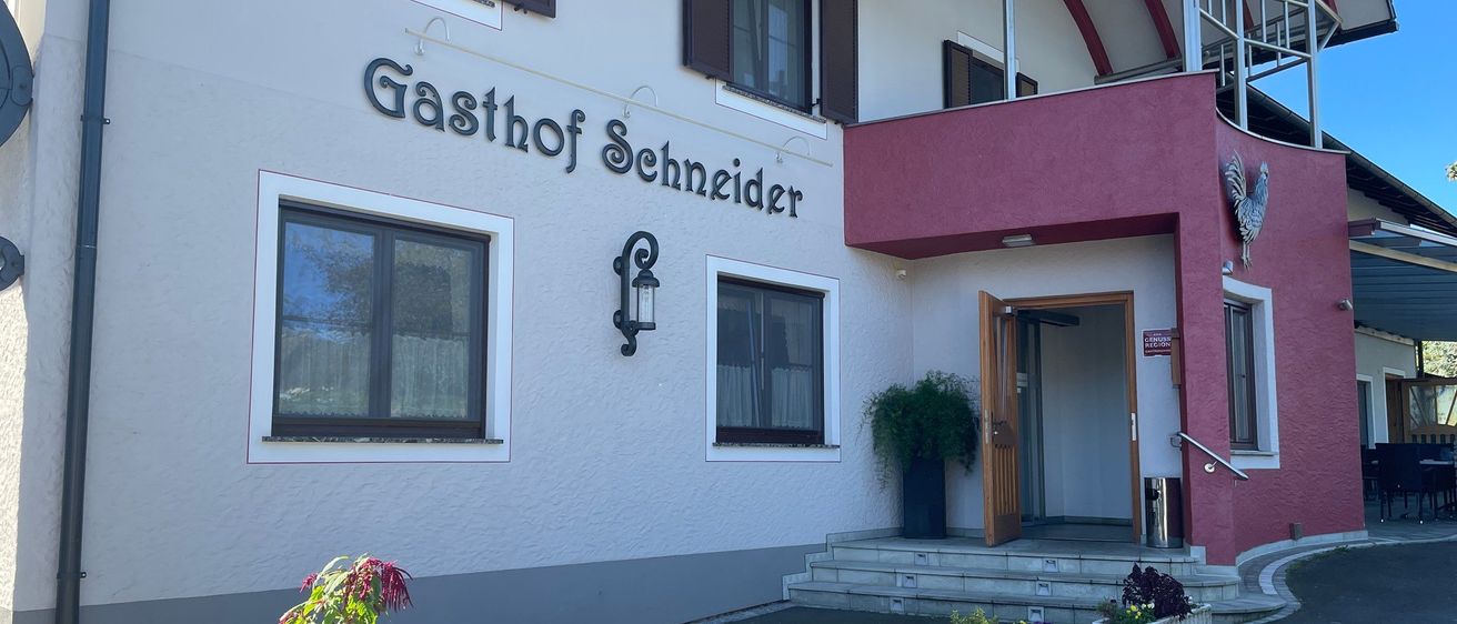 Ein weißes Gebäude mit dem Namen Gasthof Schneider. Es hat einen rot gestrichenen Bereich, einen Balkon und eine offene Tür. Der Eingang ist von Pflanzen umgeben.