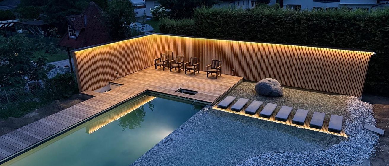 Ein Blick von oben auf einen modernen Hinterhof mit Swimmingpool und Holzdeck, beleuchtet von warmen Lichtern. Stühle und eine Lounge-Ecke umgeben den Pool, mit Felsen und Kies in der Nähe.