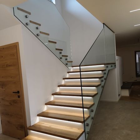 Eine moderne Treppe mit Glasstufen und Holzhandläufen, beleuchtet von weißen Lichtern. Die Treppe befindet sich in einem modernen Haus mit weißen Wänden und einer Holztür.