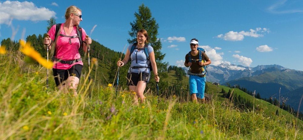 Drei Wanderer mit Rucksäcken gehen auf einem grasigen Feld mit Blick auf Berge. Einer trägt ein rosa Shirt, ein anderer einen weißen Visier und der dritte ein braunes Shirt.
