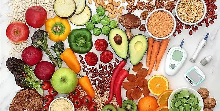 A variety of foods including fruits, vegetables, nuts, and legumes are arranged on a white marble surface. Also present are a blood glucose meter and a blood glucose test strip.
