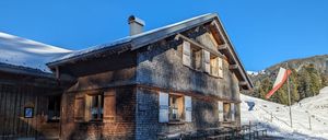 Ein Holzchalet mit schneebedecktem Dach, mehreren Fenstern und einem Schornstein. Draußen sind Tische und Bänke aufgestellt, und der Boden ist schneebedeckt.