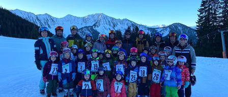 Eine Gruppe von Skifahrern in nummerierten Jacken, Helmen und Schutzbrillen posiert für ein Foto auf einem verschneiten Berg. Der Hintergrund zeigt schneebedeckte Berge und einen klaren blauen Himmel.