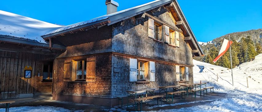 Ein Holzhaus mit Schnee auf dem Dach, umgeben von einer verschneiten Landschaft. Draußen stehen mehrere Bänke und Tische.