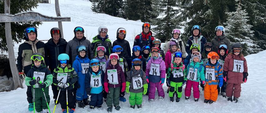 Eine Gruppe von Menschen in Skiausrüstung steht auf einem verschneiten Berg, mit Bäumen und einem Kreuz im Hintergrund. Sie umfasst Kinder und Erwachsene, einige mit Nummern auf ihren Kleidungsstücken.