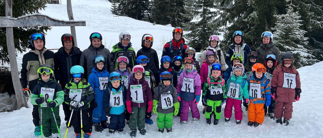 Eine Gruppe von Menschen in Skiausrüstung steht auf einem verschneiten Berg, mit Bäumen und einem Kreuz im Hintergrund. Sie umfasst Kinder und Erwachsene, einige mit Nummern auf ihren Kleidungsstücken.