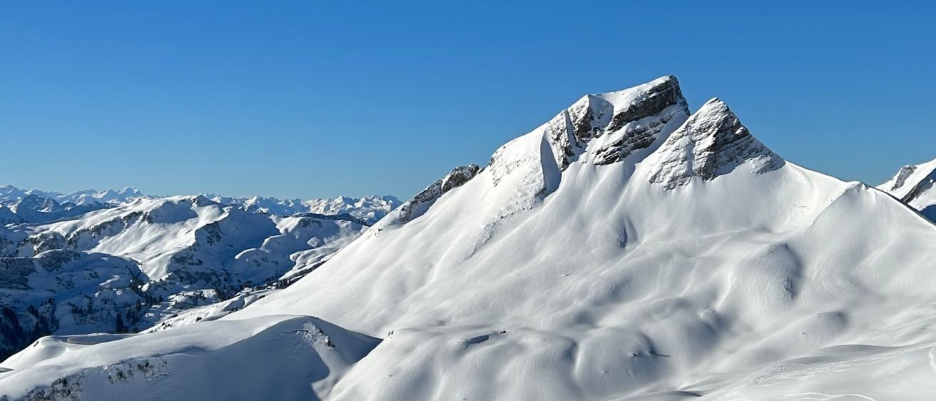 Ein Panoramablick auf eine verschneite Bergkette unter einem klaren blauen Himmel. Die Berge sind schneebedeckt und haben mehrere Skiloipen.