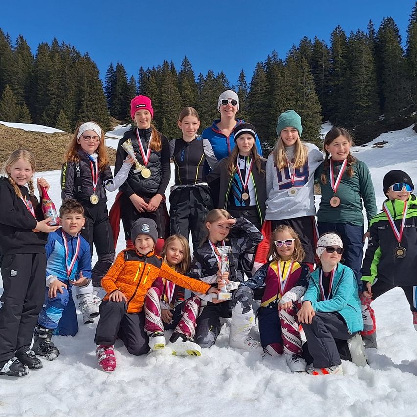 Eine Gruppe junger Skifahrer und Snowboarder, die Medaillen tragen und Pokale in der Hand halten, posiert für ein Foto im Schnee. Sie sind von Bäumen und einem klaren blauen Himmel umgeben.