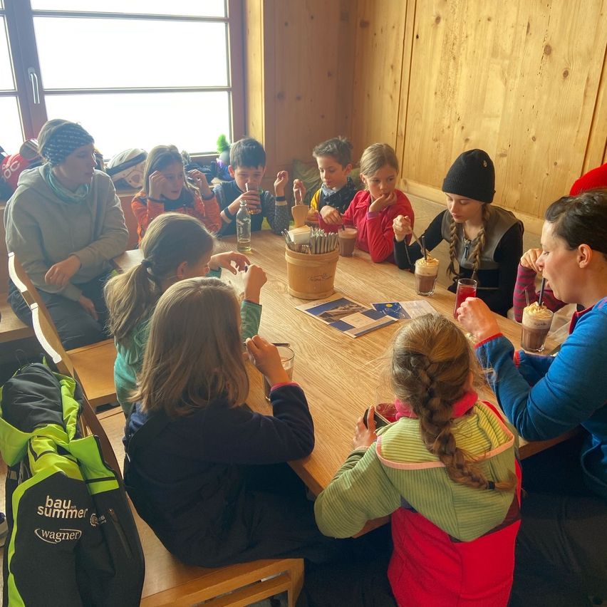 Gruppe von Kindern und Erwachsenen, die um einen Tisch im Innenbereich sitzen, einige mit Getränken in der Hand, und eine Frau mit einem Löffel.