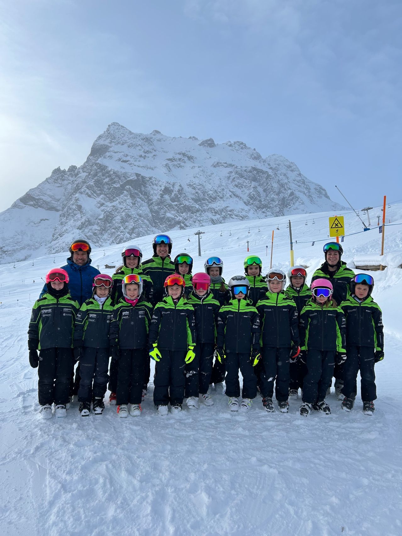 Eine Gruppe von Menschen in Skiausrüstung posiert für ein Foto auf einem verschneiten Berg. Sie tragen Helme und Schutzbrillen, mit einigen Erwachsenen hinter ihnen.