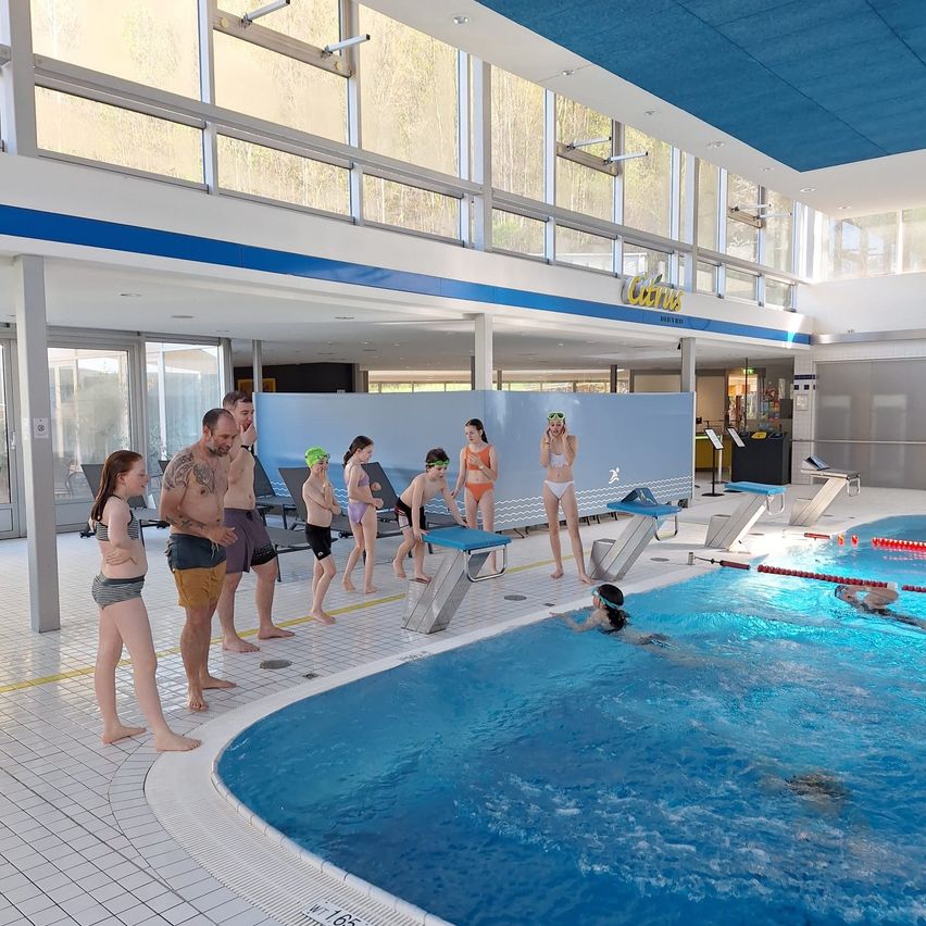 An indoor pool with swimmers and instructors, one swimmer is diving into the pool. There are glass windows on the walls.