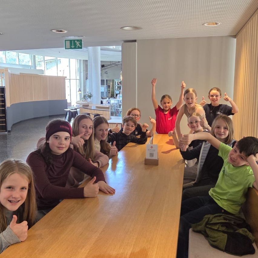 A group of young people are posing for a photo at a table in a cafeteria, some with thumbs up. Behind them, a counter and a window with an exit sign.