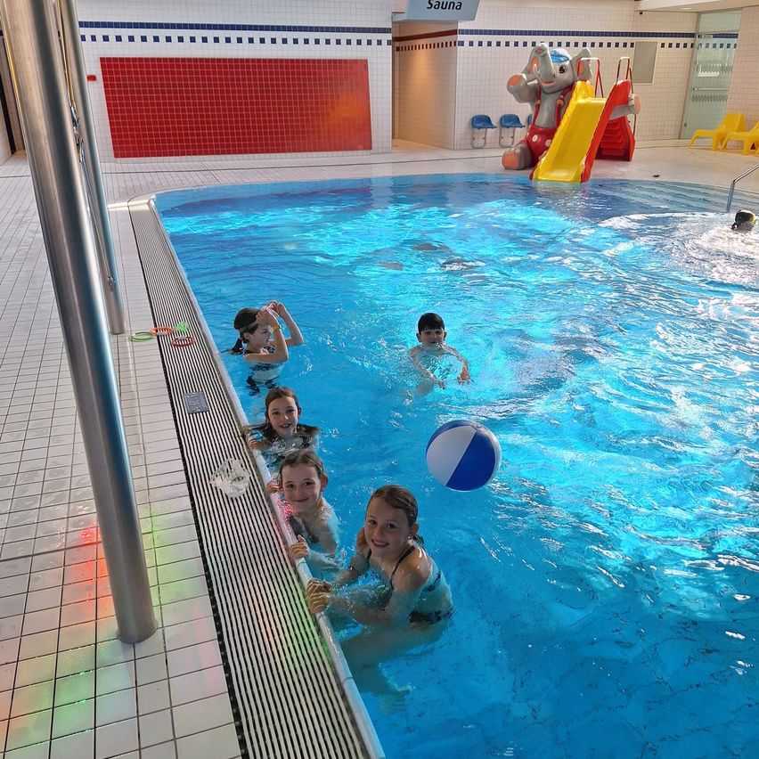 Five children are swimming in a pool, some holding a ball, while a red wall and a slide are in the background.