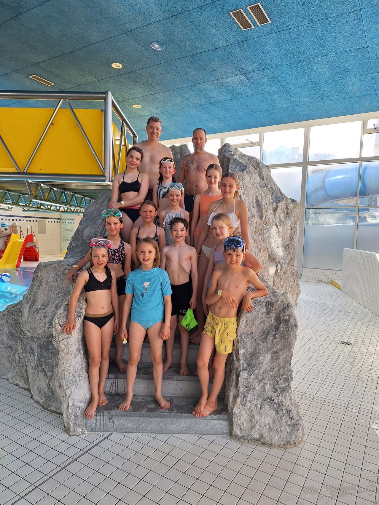 A group of children in swimwear pose for a photo in an indoor pool area, with adults standing behind them. The pool is surrounded by a rock formation.