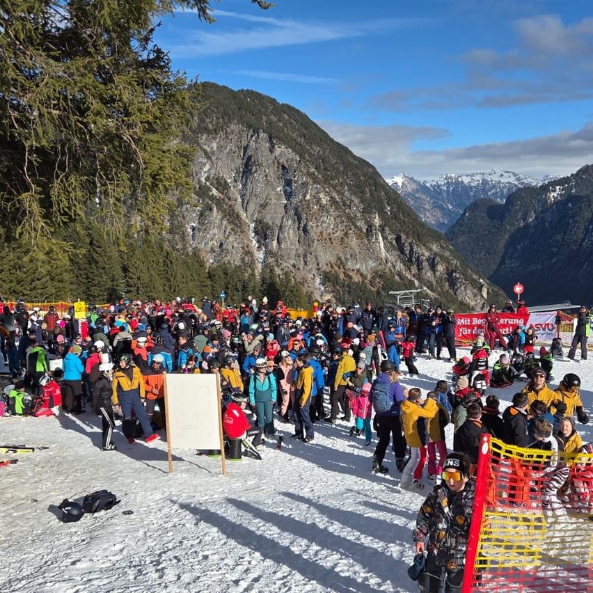 Eine große Menschenmenge versammelt sich auf einem verschneiten Berg, viele tragen Winterkleidung und Helme. Sie warten wahrscheinlich darauf, dass eine Veranstaltung beginnt.