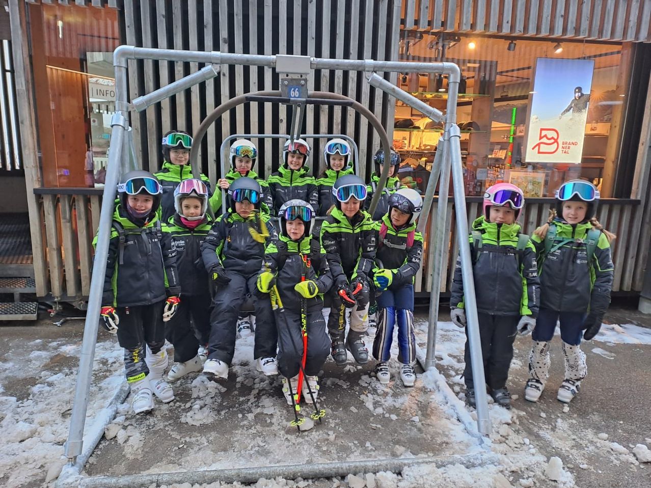 Eine Gruppe von Kindern in Skiausrüstung steht zusammen, hält Skistöcke in der Hand, mit Schnee um sie herum und einem Skilift im Hintergrund.