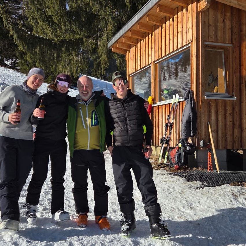 Vier Personen stehen im Schnee vor einer Holzhütte, Bierflaschen in der Hand. Sie tragen Winterkleidung und scheinen zu lächeln. Skier sind an der Hütte gelehnt.