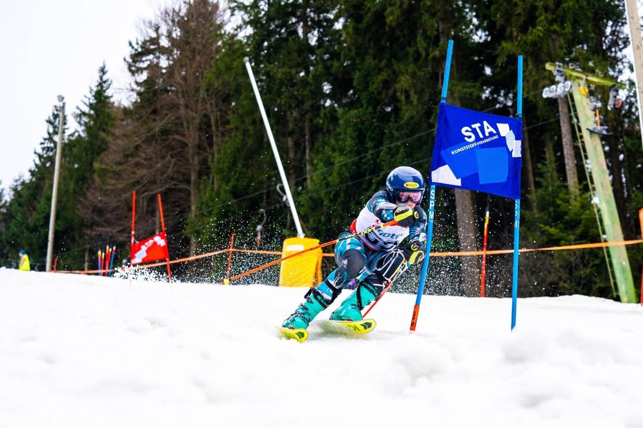 Ein Skifahrer navigiert eine Abfahrtsstrecke und fährt zwischen blauen Stangen, mit einer blauen Fahne und verschneiten Boden im Hintergrund.