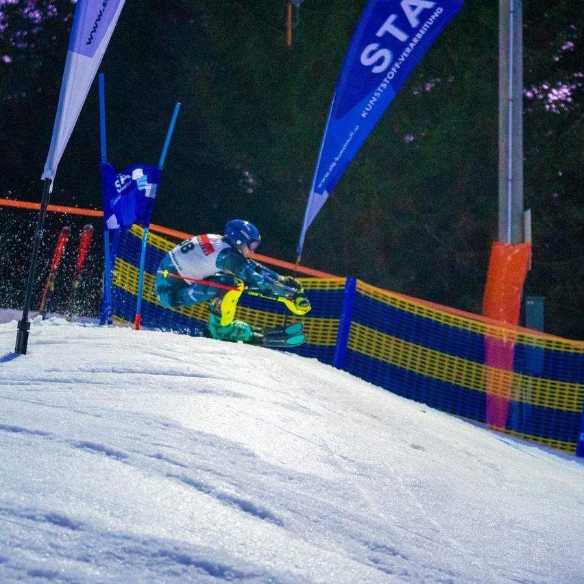 Ein Skifahrer in Helm und Skiausrüstung fährt bei Nacht einen schneebedeckten Hang hinab und passiert dabei blaue und gelbe Barrieren.