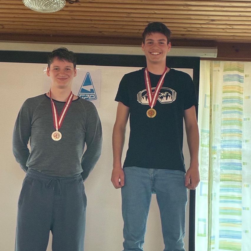 Zwei junge Männer stehen nebeneinander und tragen beide Medaillen. Sie lächeln und scheinen für ein Foto zu posieren. Hinter ihnen befindet sich eine Tafel mit einem Logo.