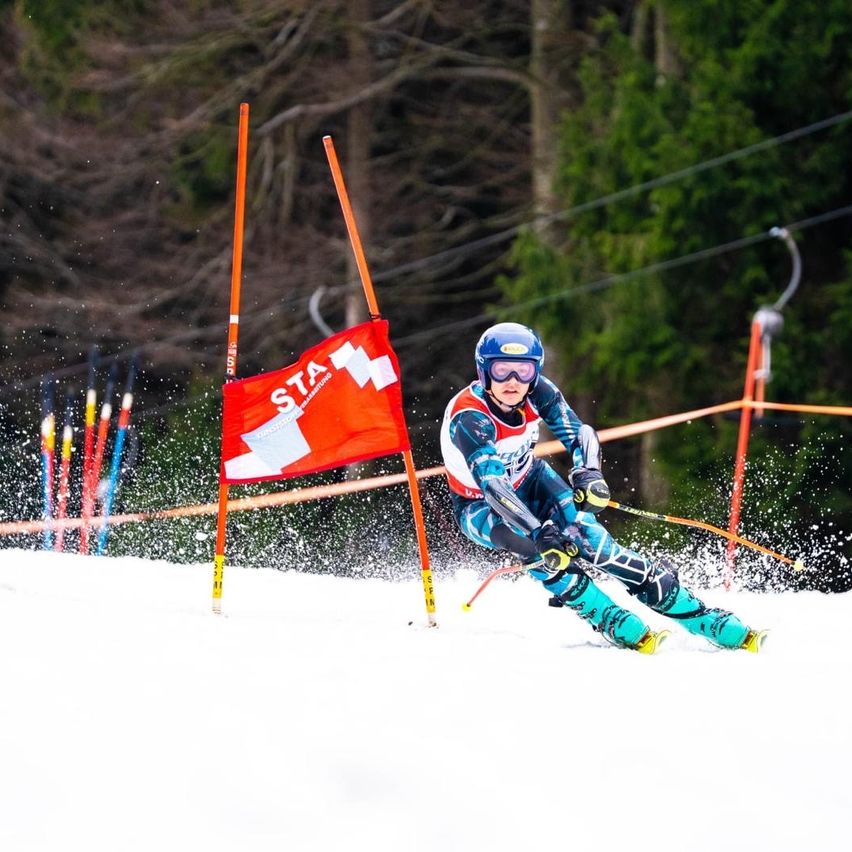 Ein Skifahrer mit blauem Helm und Schutzbrille fährt einen schneebedeckten Hang hinunter und navigiert an orangefarbenen Stangen mit einer roten Flagge vorbei.