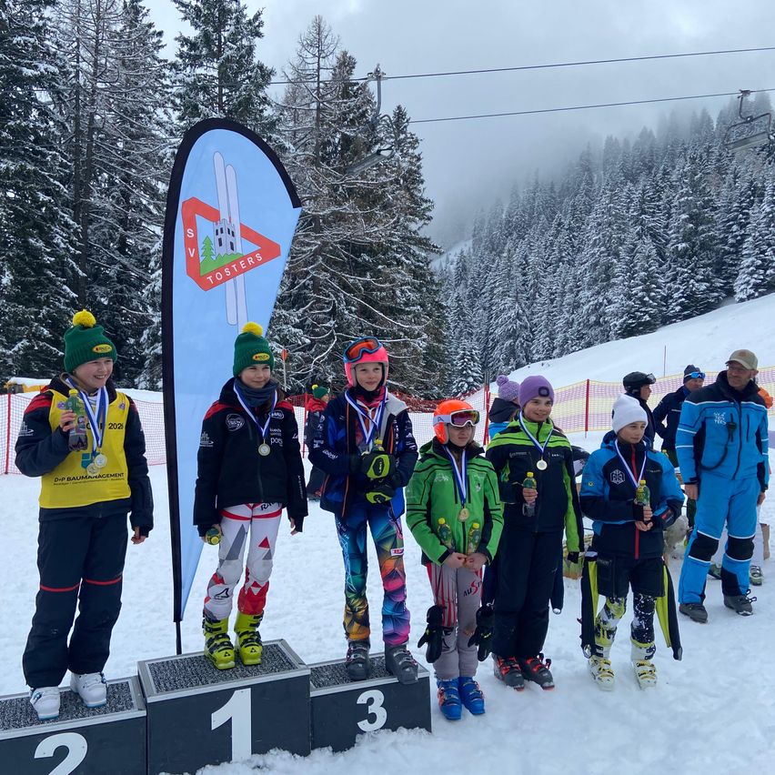 Eine Gruppe junger Skifahrer steht auf einem Podest in einem verschneiten Skigebiet. Sie tragen Medaillen und Skianzüge. Hinter ihnen steht ein Banner mit der Aufschrift SV Tosters.