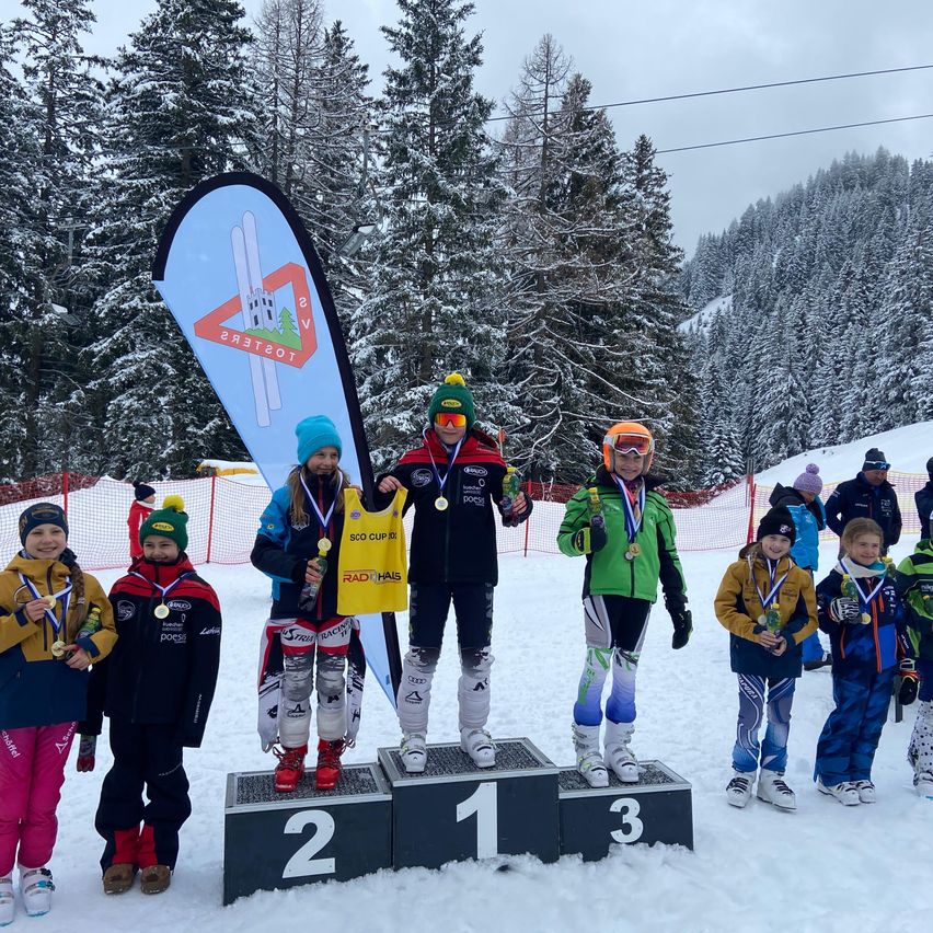 Kinder stehen auf einem Podium mit schneebedeckten Bäumen im Hintergrund. Sie tragen Winterkleidung und halten Medaillen. Drei sind auf dem Podium, und andere sind in der Nähe. Eine Person hält ein gelbes Banner.