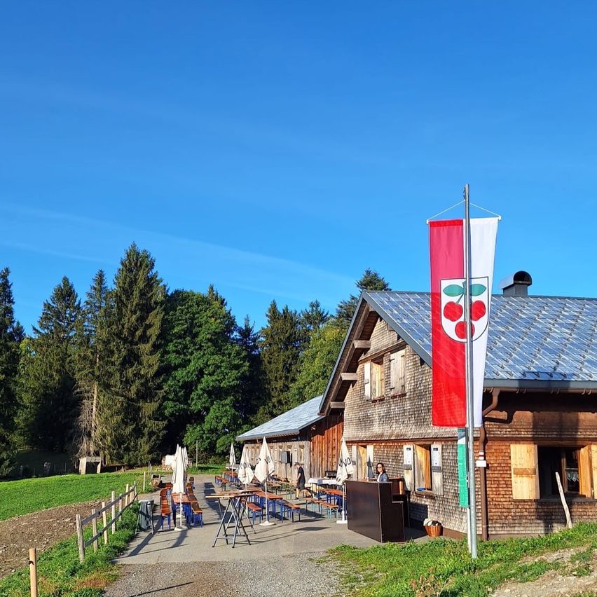 Ein rustikales Holzgebäude mit einer rot-weißen Fahne auf einem Pfosten steht an einem Hang mit blauem Himmel im Hintergrund.