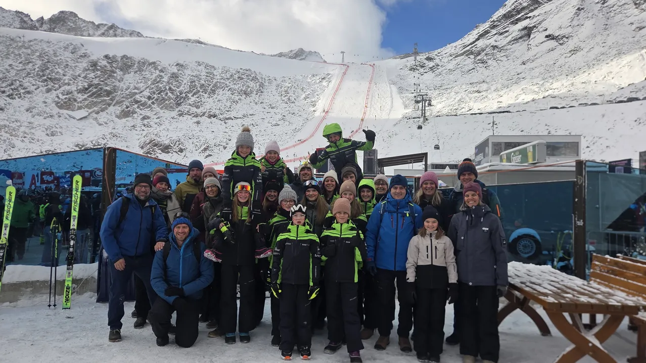Eine Gruppe von Menschen posiert für ein Foto in einem verschneiten Berggebiet. Sie tragen alle Winterkleidung und einige von ihnen haben Skier an den Füßen.