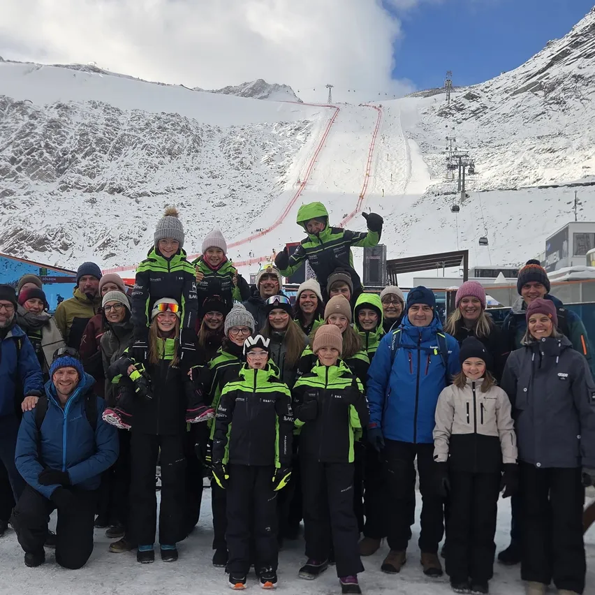 Eine Gruppe von Menschen posiert für ein Foto in einem verschneiten Berggebiet. Sie tragen alle Winterkleidung und einige von ihnen haben Skier an den Füßen.