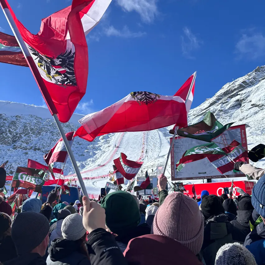 Eine Menschenmenge versammelt sich bei einem Ski-Event und schwenkt Flaggen. Die Flaggen sind überwiegend rot und weiß mit einem Adleremblem. Die Veranstaltung findet in einem Gebirgsgebiet mit klarem blauem Himmel statt.