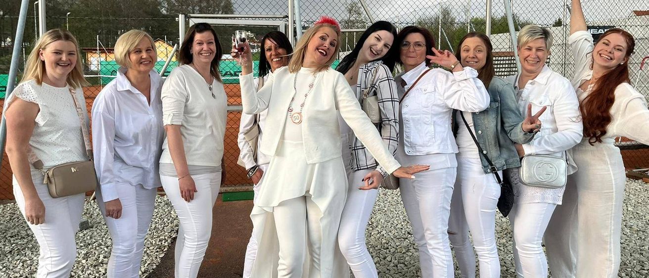 Eine Gruppe von Frauen in passenden weißen Outfits posiert für ein Foto vor einem Zaun mit bewölktem Himmel im Hintergrund.