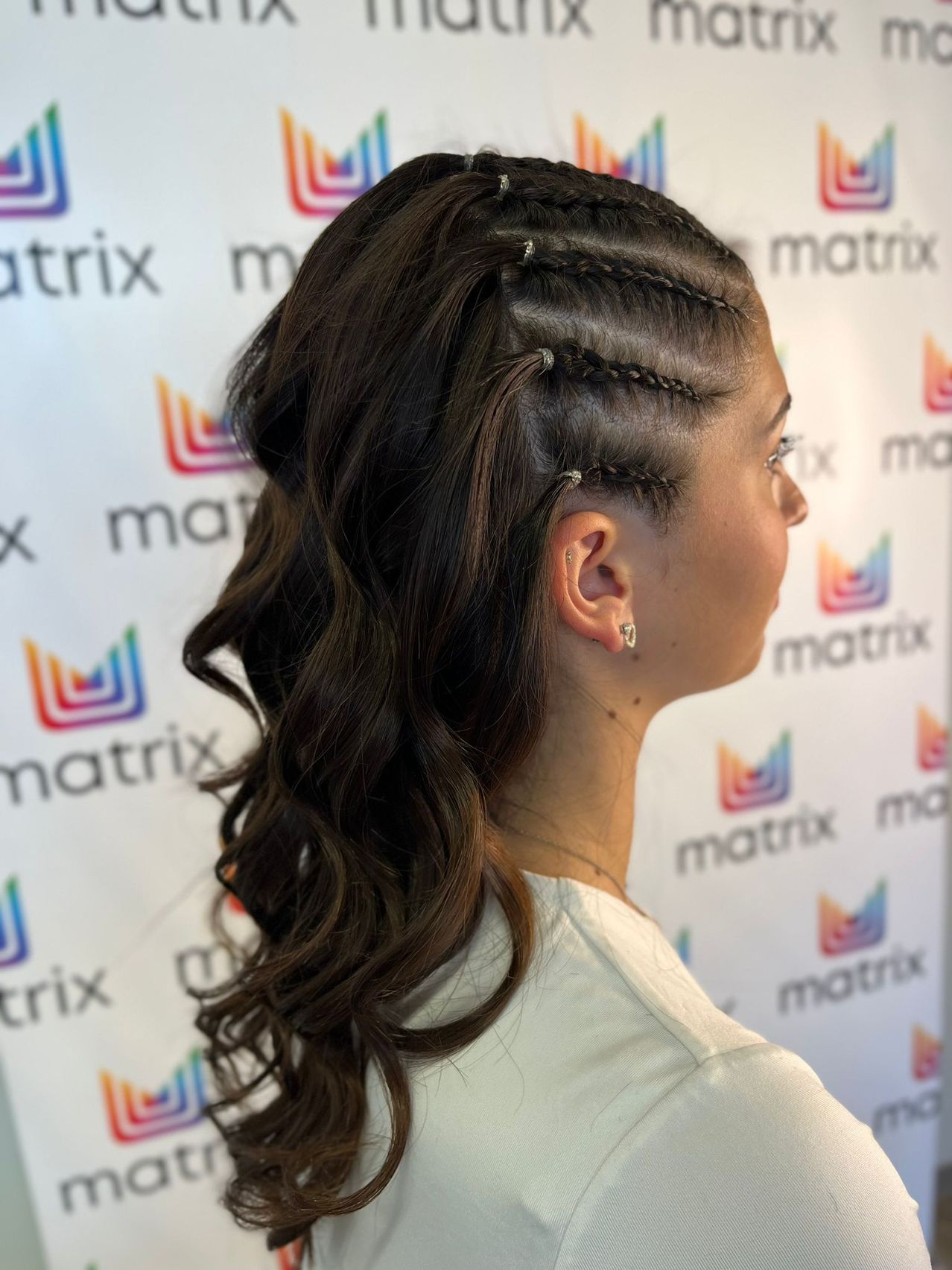 Eine Frau mit langen, dunklen Haaren, kunstvoll geflochten, steht vor einem weißen Hintergrund mit bunten Matrix-Logos. Sie trägt ein weißes Shirt und ihr Profil ist sichtbar.