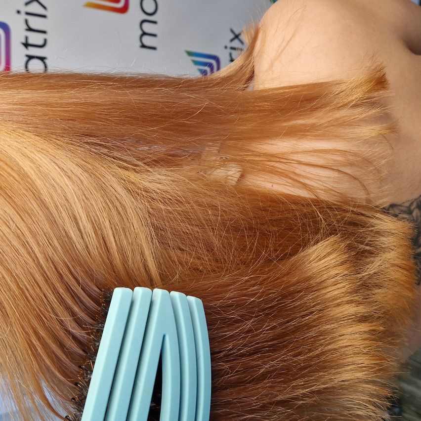 Eine Nahaufnahme von jemandem, der sein langes, braunes Haar mit einer türkisen Haarbürste bürstet. Im Hintergrund ist ein weißes Schild mit blauen und grünen Logos zu sehen.
