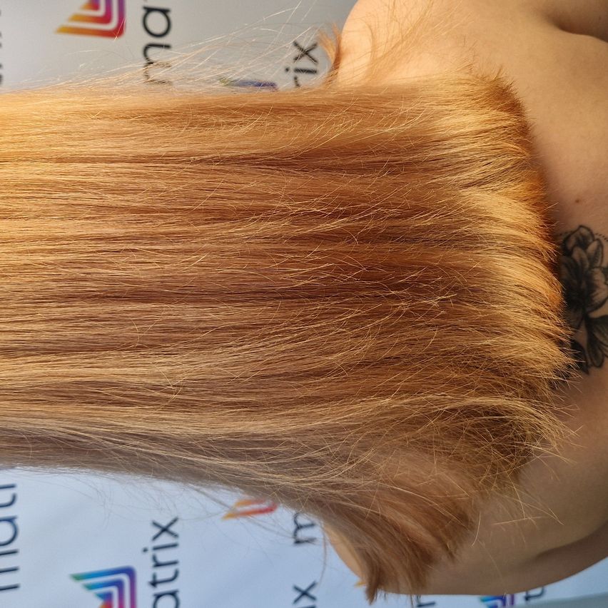 Eine Nahaufnahme der Schulter einer Person mit hellbraunem Haar. Das Haar ist leicht gewellt und hat einen natürlichen Glanz. Ein Tattoo einer Blume ist auf der Schulter sichtbar.