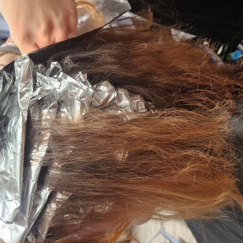 Eine Hand eines Menschen ist zu sehen, wie sie Folie auf braunes Haar aufträgt, wahrscheinlich zum Färben oder Hervorheben. Die Folie ist silberfarben und um das Haar gewickelt.