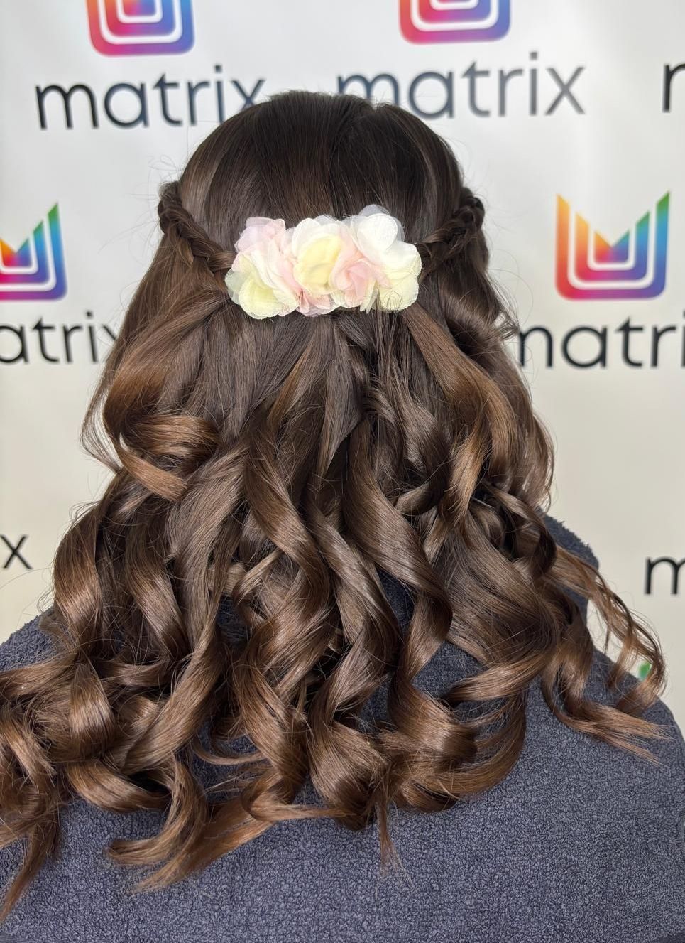 Ein Blick von hinten einer Frau mit lockigem braunem Haar, geschmückt mit einer rosa-weißen Blumenhaarnadel.