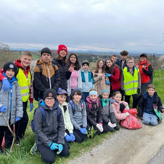 Gruppe von Kindern und Jugendlichen mit Handschuhen und Warnwesten, einige halten Pflanzen, lächeln für ein Foto an einer grasigen Straßenseite mit einer malerischen Aussicht im Hintergrund.