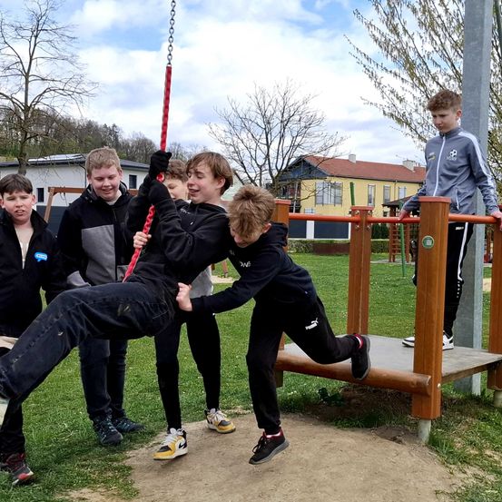 Fünf junge Jungen spielen auf einem Spielplatz; ein Junge auf einer Schaukel, ein anderer hält die Schaukel fest, während der dritte Junge versucht, sein Bein zu greifen.